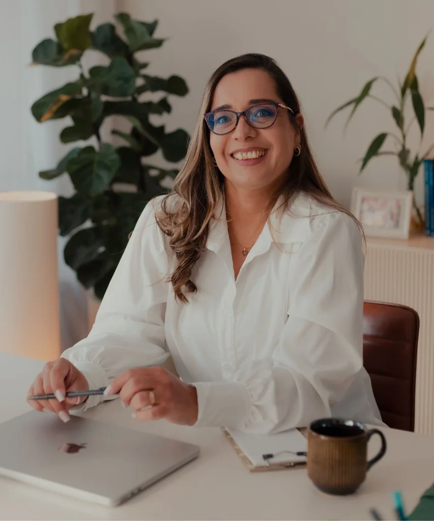 Ary Aguilar sorrindo, sentada à mesa com um caderno e uma caneta.